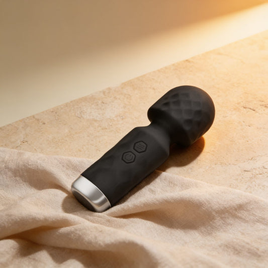 Mini wand en silicone noir — posé sur du lin drapé, lumière chaude éditoriale